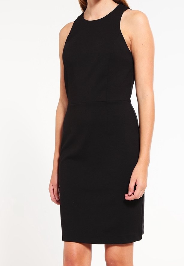 Femme portant une robe noire sans manches, longueur genou, avec un col rond sur un fond blanc uni.