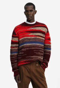 Strickpullover mit kräftigen roten, blauen und goldenen Querstreifen, einem Rundhalsausschnitt und strukturierten Oberflächen, kombiniert mit braunen Hosen.