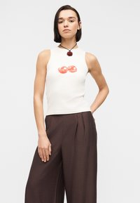 Femme portant un haut blanc sans manches avec un motif de cerise rouge et un pantalon marron à jambes larges, debout avec une main dans la poche, sur fond blanc.