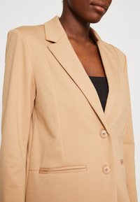 Beige skreddersydd blazer med snappelapper, to knapper og diskret frontlomme-detaljering i glatt stoff, som delvis dekker en svart topp.
