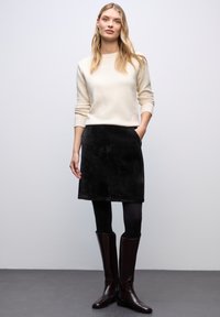 Suéter crema, falda de pana negra, medias negras y botas de caña alta marrones. La falda tiene bolsillos y una silueta recta.