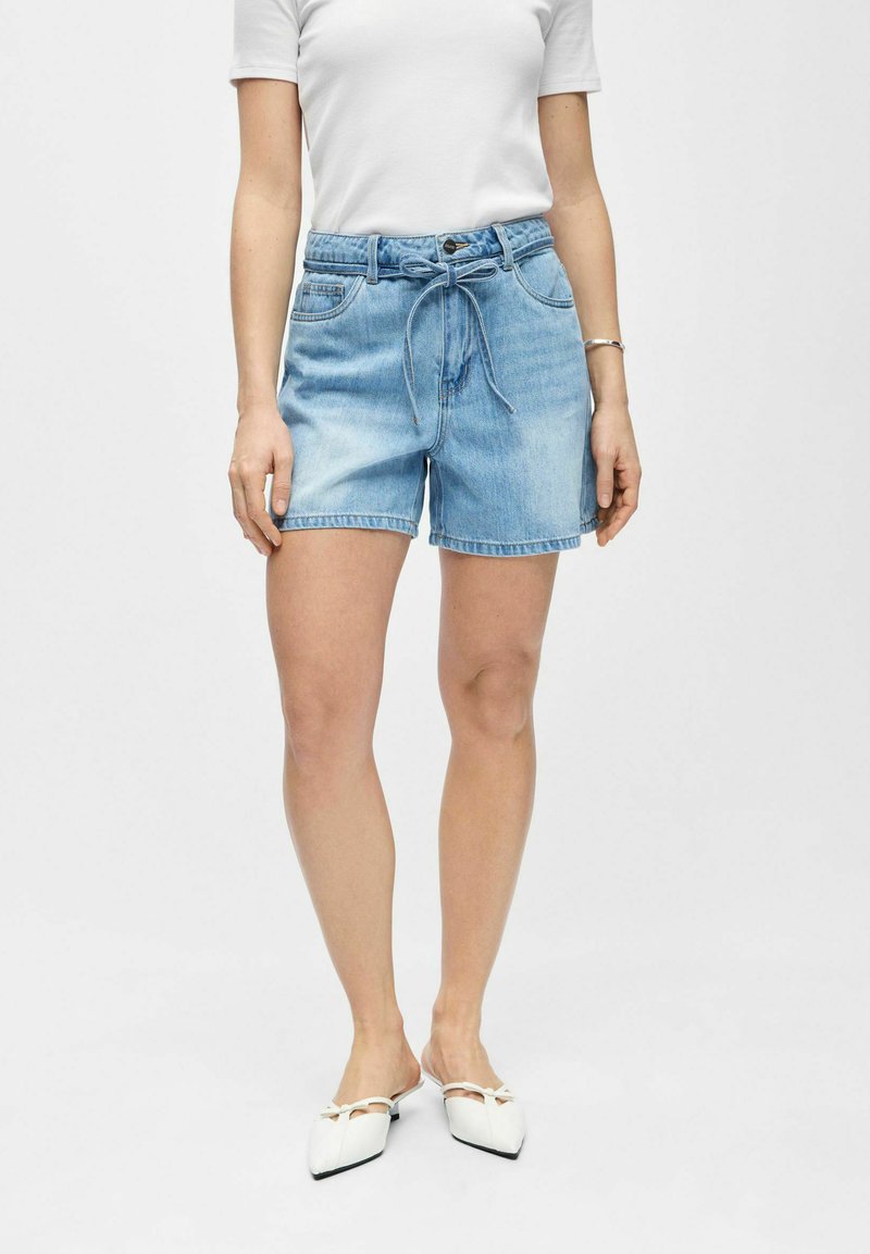 Femme portant un short en jean bleu clair avec une ceinture nouée à la taille, un t-shirt blanc et des chaussures plates blanches à bouts pointus avec des nœuds.
