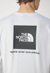 Hvid langærmet t-shirt med et sort rektangulært logo med teksten "THE NORTH FACE" og "NEVER STOP EXPLORING" på ryggen.