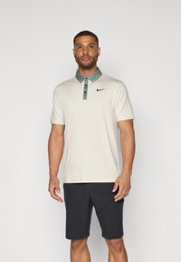 Beige polo krekls ar zaļu apkakli, trīs pogu aizdari, melnu Nike logo, salikts ar tumšām šortiem. Mīksts audums un klasiskā dizains.