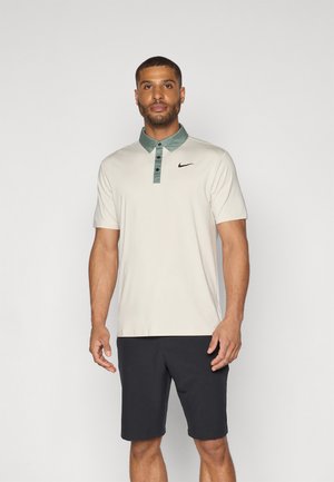 Beige poloshirt met een groene kraag, drie-knopen pat, zwart Nike-logo, gecombineerd met donkere shorts. Zachte stof en klassiek ontwerp.
