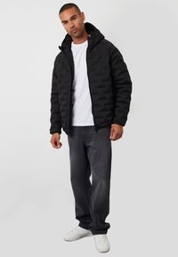 Zwarte gewatteerde jas met capuchon, met een gestikte ontwerp. Draag het over een witte T-shirt, gecombineerd met donkergrijze jeans en witte sneakers.