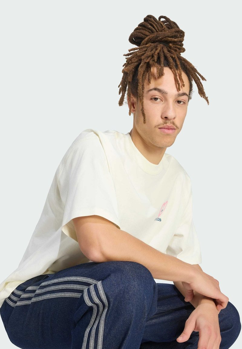 Junge Person mit langen Dreadlocks, zu einem Knoten gebunden, trägt ein weißes, übergroßes T-Shirt und blaue Jogginghosen mit weißen Streifen, sitzt mit einem angewinkelten Knie.