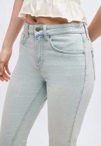 Heleõrnad helerohelised kõrge vöökohaga teksapüksid, millel on nööp- ja tõmblukuga kinnitamine, kantud koos lühikese valge topiga, mis paljastab osa vööst ja kätt.