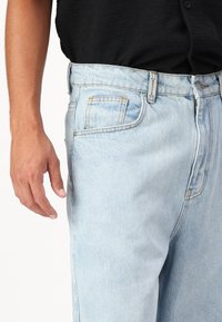 Jeans de denim azul claro con corte relajado, que cuentan con cinco bolsillos, costuras amarillas y un cierre de botón plateado. La tela tiene una textura ligeramente marcada.