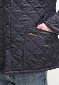 Könnyű, steppelt sötétkék dzseki patentgombos záródással, átlós varrással és narancssárga hímzett "Barbour" logóval az elülső zsebén.