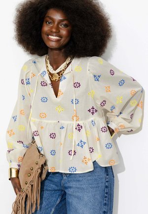 Femme souriante avec un afro volumineux porte un chemisier crème avec des motifs brodés colorés, des colliers dorés superposés, un jean bleu et porte un sac à franges beige.