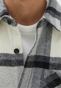 Gray and white plaid flannel shirt with button closure, black buttons, and pocket detail. White ribbed crew neck shirt underneath.