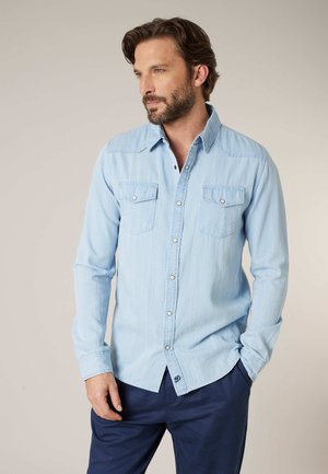 Man met baard die een lichtblauw overhemd met knopen en een donkerblauwe broek draagt, staat met één hand in zijn zak en kijkt opzij.