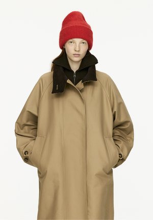 Manteau oversize beige avec un col marron, doté de poches latérales et de détails en boutons. Un bonnet en tricot rouge sur la tête ajoute un contraste.