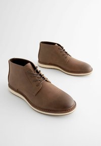 Botas de cuero marrón hasta el tobillo con punta redonda, sistema de cordones y suela de color crema. Textura suave con detalles de costura en contraste.