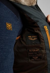Tissu texturé bleu marine à gauche, avec un patch logo orange. Le tissu extérieur vert foncé est rehaussé par une fermeture éclair orange vif et des étiquettes de doublure marron.