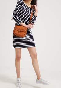 Marineblaue und weiße, gestreifte Langarmkleid, kombiniert mit einer beigefarbenen Umhängetasche aus Leder mit perforierten Details und braunen Trägern. Silberfarbene Sneaker.