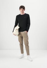 Schwarzes Langarmshirt mit einem Logo, beigefarbene schmal zulaufende Hose und weiße Sneakers, hält eine cremefarbene Tasche mit Struktur und Falten.