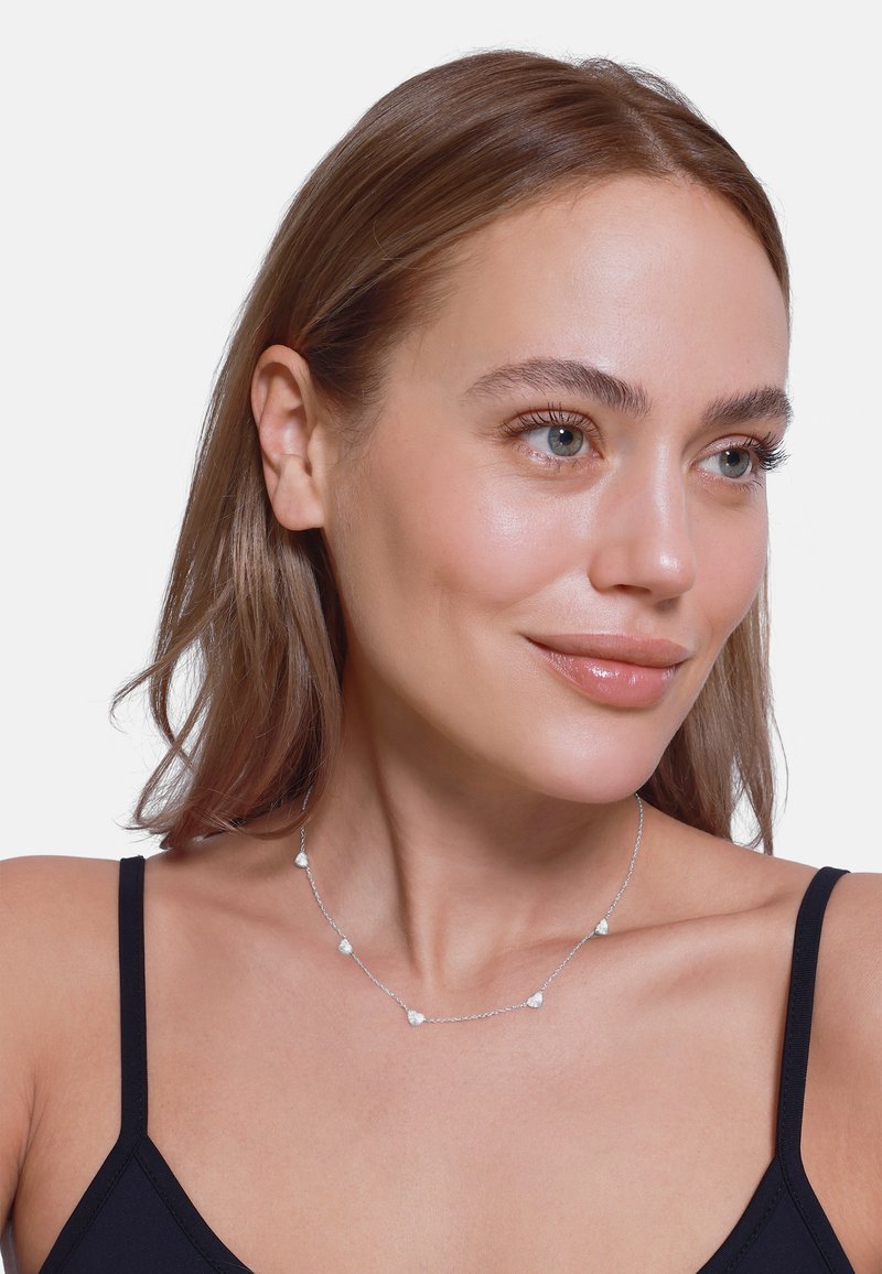 Collana d'argento con cinque accentuazioni a forma di cuore, texture liscia, design a catena sottile, indossata da un modello con capelli castani lisci.