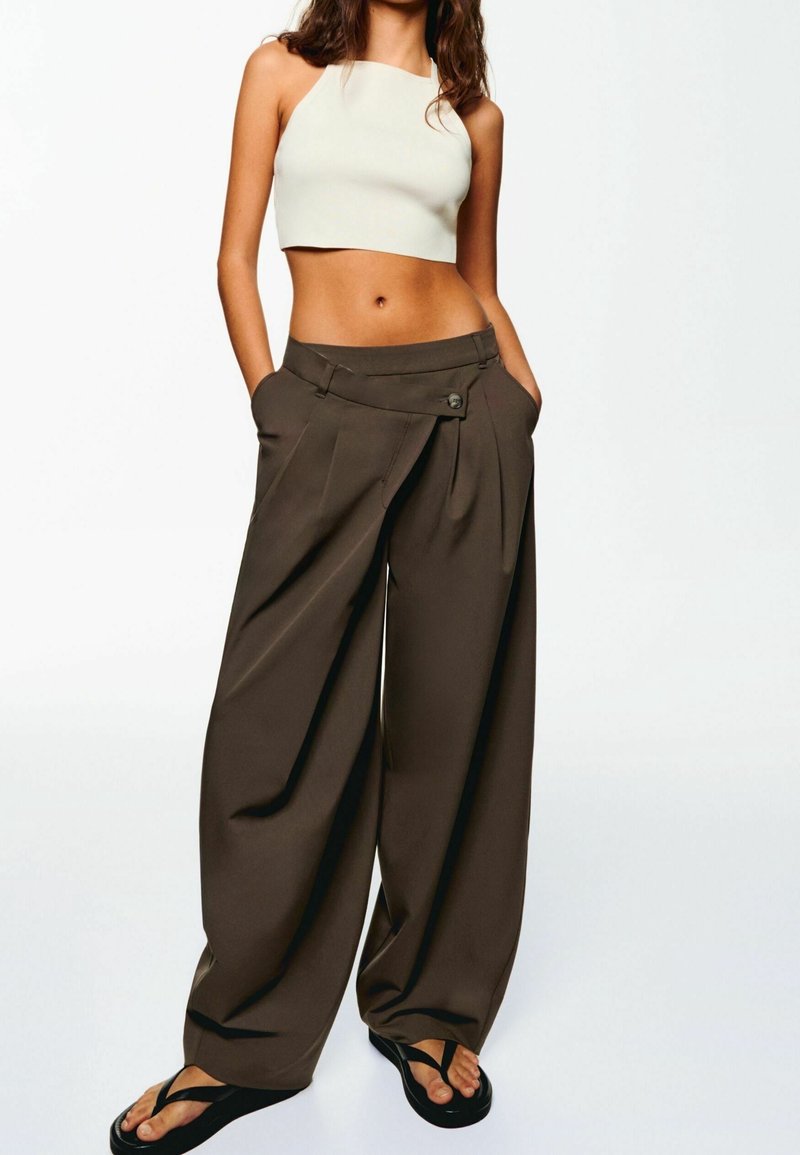 Femme portant un crop top blanc sans manches, un pantalon ample à taille haute large marron foncé, et des tongs noires, debout les mains dans les poches.