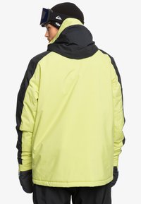 Höghalset skidjacka i starkt gult med svarta accenter, med en huva, långa ärmar och en slank, strömlinjeformad design.