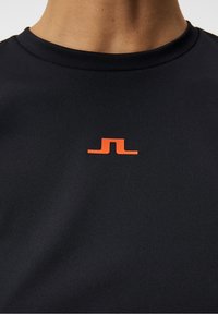 Camiseta deportiva negra de tela texturizada, con un pequeño logo naranja en el pecho. Cuello redondo y mangas cortas.