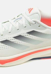Scarpa sportiva grigia chiara con upper in rete, dettagli a strisce nere, tallone arancione e suola ammortizzata con finitura testurizzata.