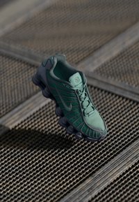 Zapatilla verde con parte superior de malla y suela de goma, que presenta un diseño texturizado y un detalle de amortiguación prominente cerca del talón.