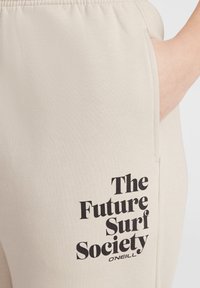 Beige sweatpants van zacht materiaal met een effen ontwerp. Opvallende zwarte tekst: "The Future Surf Society" dicht bij de zak.