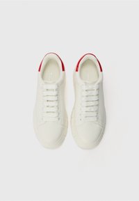 Białe sneakersy o gładkim skórzanym wykończeniu, okrągłym nosie, płaskiej podeszwie i czerwonych akcentach na pięcie, z sześcioma parami białych oczek.
