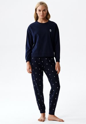 Młoda kobieta ubrana w granatową bluzkę z długim rękawem z napisem "ATLANTIC" oraz dopasowane granatowe spodnie z małym białym wzorem, stoi boso.