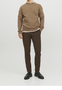Hnědá mikina s kulatým výstřihem, přes kterou je vrstvená bílá kostkovaná košile. Hnědé elegantní kalhoty a černé mokasíny doplňují celý outfit.