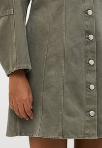 Ελαιόχρωμο τζιν φόρεμα με μακριά μανίκια, έντονες ραφές και ασημένια κουμπιά μπροστά. Διαθέτει ευθεία σταυρωτή ραφή και καθορισμένα ραφ�άκια.