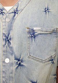 Heleheline teksavest helesinise värvusega, millel on tähte kujutav õmblustehnoloogia, eesosas tasku ja hõbedased nupud. Sildil on tekst "Ära raiska kultuuri."