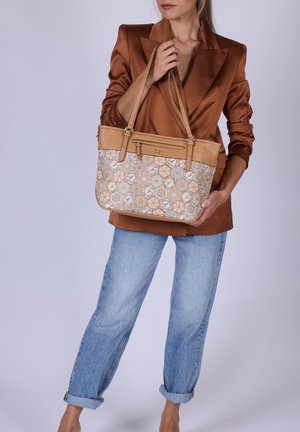 Donna con blazer di raso marrone e jeans blu risvoltati che tiene una borsa beige con motivo e spalline lunghe.