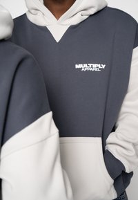 Hanorac gri și crem, cu decolteu în V, buzunar mare în față și logo "MULTIPLY APPAREL" alb. Material moale cu culori contrastante.