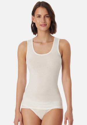 Vrouw met kort bruin haar, gekleed in een nauwsluitend wit tanktopje en bijpassend wit ondergoed, staand tegen een effen witte achtergrond.