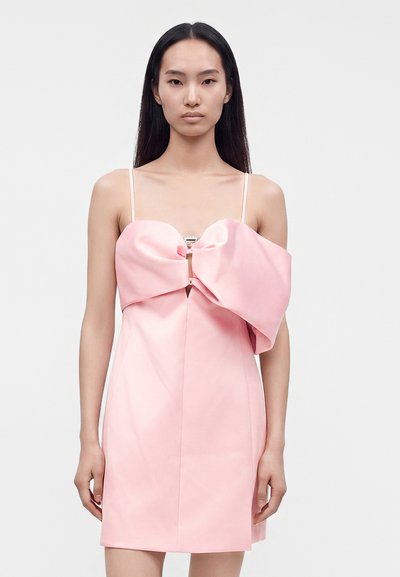 Jeune femme aux longs cheveux noirs portant une mini-robe rose clair sans manches avec un grand nœud sur la poitrine.