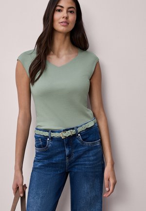 Femme portant un haut sans manches à encolure en V vert clair, rentré dans un jean bleu avec une ceinture tressée assortie, tenant une poignée marron.