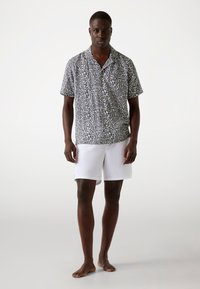 Short-sleeve black and white patterned shirt with buttons and white swim shorts. Model stands barefoot against a light background.
