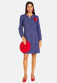 Blau gemustertes Kleid mit langen Ärmeln, einem Kragen und roten Akzenten. Das Model hält einen runden roten Hut, trägt rote Absätze und kreisförmige Ohrringe.