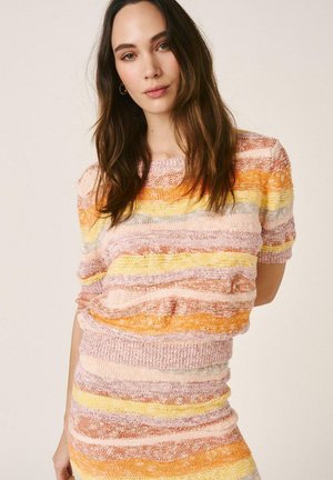 Joven con cabello largo y castaño, vestida con un top de punto a rayas pastel y falda a juego, posando contra un fondo liso.