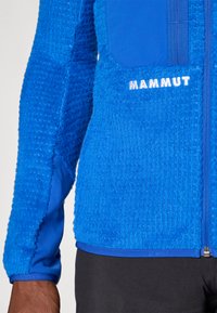 Μπλε φλις μπουφάν με υφή, λείες πλευρικές πάνελ, κλείσιμο με φερμουάρ και λογότυπο "Mammut" σε άσπρο μπροστά.