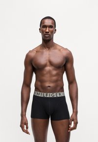 Črne bokserice z sivo elastično pasom, na katerem je v črnem tisku napis "HILFIGER". Tkanina se zdi gladka in raztegljiva. Model pozira spredaj.