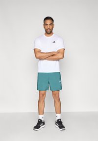 Fehér rövid ujjú sportpóló fekete logóval, hozzá illő türkiz rövidnadrág logóval. Fekete sportcipő, fehér zokni.