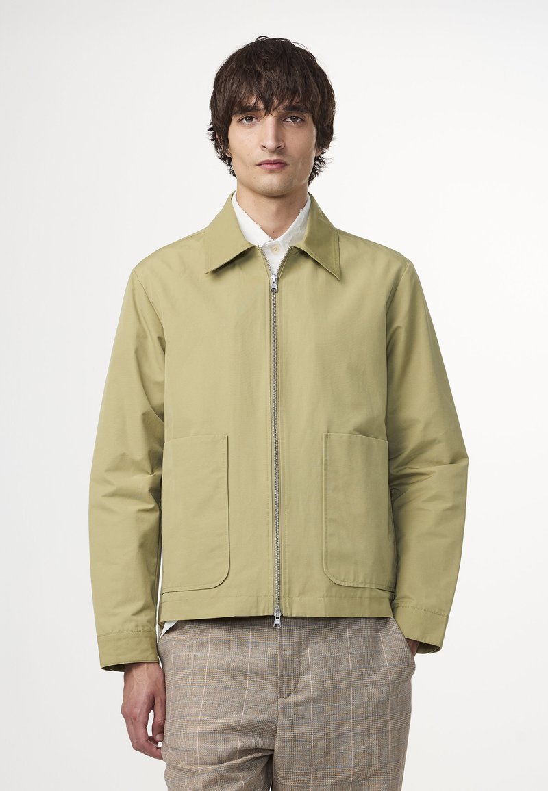 Ljusgrön zip-up jacka med klassisk krage, två framfickor och mjuk textur, bärs över en vit skjorta och mönstrade byxor.