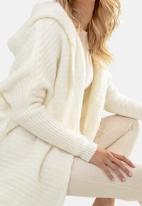 Cardigan bianco lavorato a maglia con cappuccio, con una texture a costine sulle maniche e sul corpo. Indossato sopra un top chiaro e abbinato a pantaloni crema.