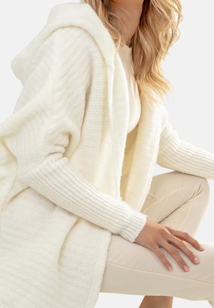 Vit stickad cardigan med huva, med ribbad struktur på ärmar och kropp. Bärs över en ljus topp och kombineras med krämfärgade byxor.