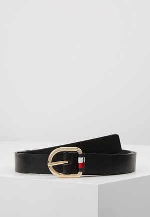 Ceinture en cuir noir avec boucle rectangulaire dorée aux bords arrondis et petit détail en tissu rouge, blanc et bleu près de la boucle, présentée sur une surface blanche.