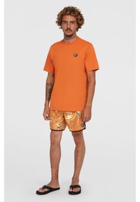 Orange bomulds t-shirt, sort logo patch, parret med knælange orange shorts der har et virvlende mønster i sort og fløde, sorte sandaler.
